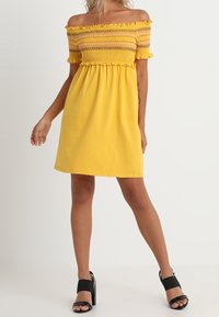 Žluté šaty s odhalenými rameny, s elastickým smocked živůtkem, volánkovými rukávy a uvolněným střihem, doplněné černými sandály na blokovém podpatku.