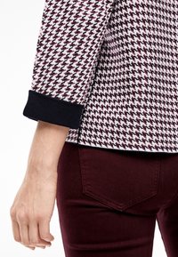 Haut à manches longues en motif à carreaux noir, blanc et bordeaux ; doté de poignets en tricot. Associé à un pantalon bordeaux, mettant en valeur une texture lisse.