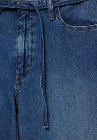 Prim-plan al taliei blugilor din denim albastru, cu bucle pentru curea, buzunar frontal și o curea din același material legată lejer.