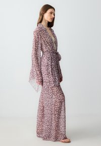 Robe en imprimé léopard transparent avec des manches longues, taille ceinturée et silhouette fluide. Principalement rose avec des taches marron, en tissu léger.
