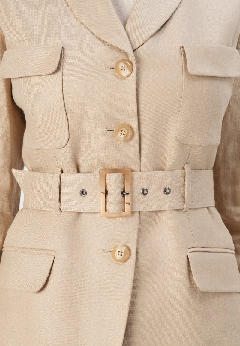 Beige maßgeschneiderte Jacke mit tailliertem Schnitt, einem Gurt in der Taille, zwei Brusttaschen und einem Reverskragen. Mit tanfarbenen Knöpfen und einem glatten Stofftextur.