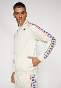 Krema zip-up trenirka s rdečimi in modrimi stranskimi črtami, ki vsebuje logotip, rebraste manšete in gladko teksturo tkanine.