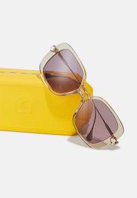 Gafas de sol de gran tamaño con tintado marrón y montura color beige claro, con un adorno decorativo de perlas, junto a un estuche texturizado amarillo.
