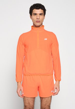 Mann steht mit dem Gesicht nach vorne und trägt ein leuchtend orangefarbenes, langärmliges Sportoberteil mit Viertelreißverschluss und passende Shorts vor einem einfachen hellen Hintergrund.