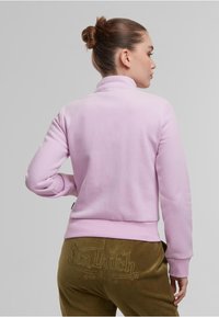Sweatshirt rose clair avec un col haut et des poignets côtelés, porté sur un pantalon vert olive avec un logo texturé. Texture de tissu lisse.