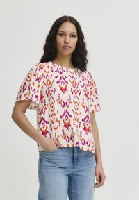 Short-sleeve top in light fabric featuring a pink, orange, and purple abstract pattern. Crew neckline and relaxed fit. Worn with jeans.