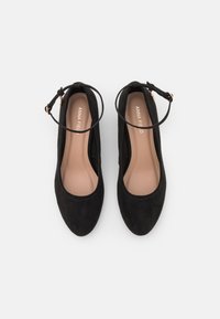 Zapatos de tacón alto de ante negro con puntera redonda, tira en el tobillo y forro interior beige en contraste. Textura suave con un detalle de costura mínima.