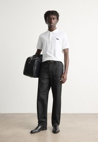 Weißes Poloshirt, dunkle Jeanshose, schwarze Lederstiefel und eine schwarze Reisetasche. Der Modell steht vor einem hellen Hintergrund mit minimalen Details.