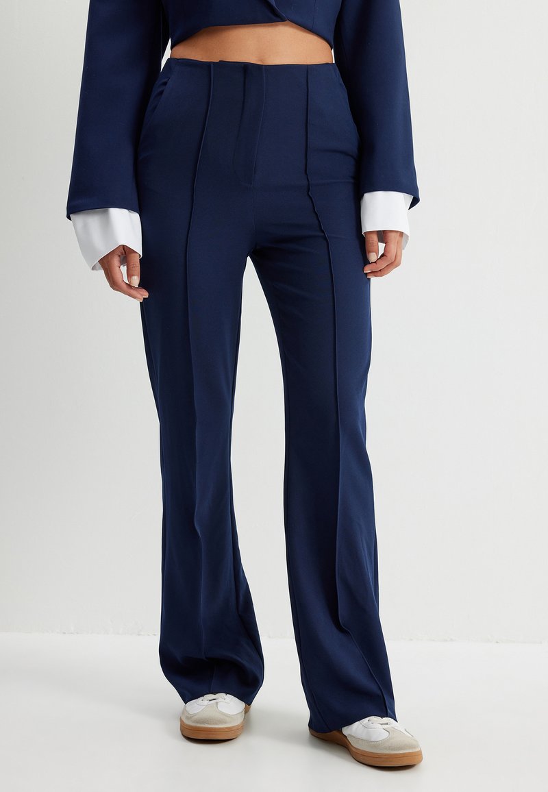 Pantalons taille haute bleu marine avec un design évasé, dotés de plis avant et d'une texture de tissu lisse, associés à des manches blanches à revers.