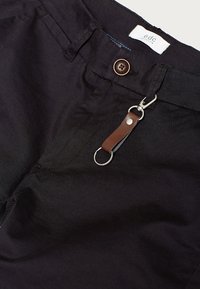 Pantaloni neri in tessuto con un'etichetta in pelle marrone, accessori in argento, chiusura a bottone e tasche laterali con una consistenza liscia.
