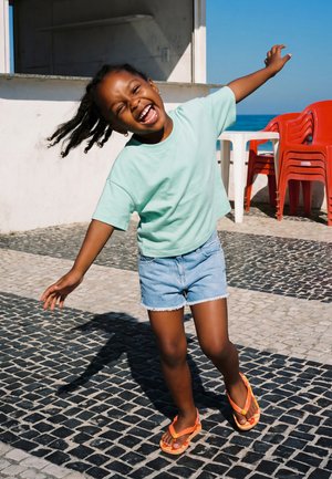 Young girl in light green shirt and denim shorts joyfully balancing on one foot on a tiled outdoor patio near red chairs and ocean.