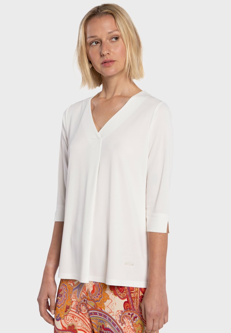 Blusa blanca de cuello en V con mangas tres cuartos, hecha de tela suave. Presenta costuras sutiles y un ajuste relajado, combinada con pantalones estampados.