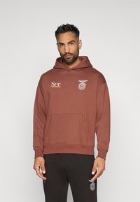 Brązowa bluza z kapturem z kieszenią z przodu, z białymi i złotymi haftowanymi akcentami. Noszona z czarnymi spodniami dresowymi z logo.