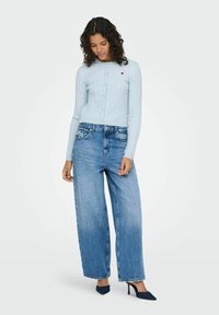 Gaiši zils adīts kardigans ar sirds motīvu, kopā ar vaļīgām zilām džinsa biksēm un tumši zilām augstpapēžu kurpēm.