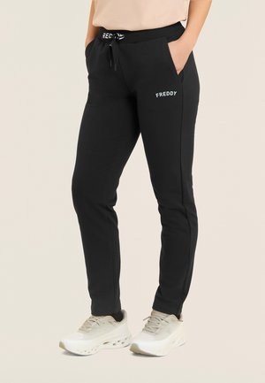 Persona che indossa pantaloni jogger neri del marchio Freddy con tasche laterali e sneakers beige, con le mani nelle tasche, in piedi su sfondo chiaro.