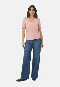Top corto de manga corta de canalé color rosa claro con escote en V y detalles de encaje, combinado con unos jeans de mezclilla azul de pierna ancha. La modelo está de pie con las manos entrelazadas.