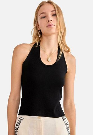 Joven con cabello largo y rubio, vestida con top sin mangas negro de canalé, pantalones blancos con bordado de hojas negras, collar con colgante de perlas y pendientes.