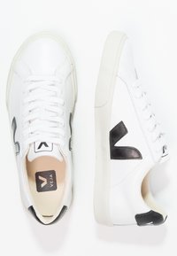 White sneakers with a smooth leather upper, featuring black accents, round toe, flat laces, and a textured rubber sole.