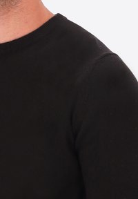 Uomo che indossa un maglione nero a girocollo, che mostra collo, spalla e parte superiore del braccio su uno sfondo chiaro e neutro.