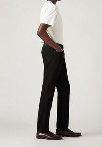 Vue latérale d'une personne portant un T-shirt blanc, un pantalon noir droit et des mocassins en cuir rouge foncé, debout les mains dans les poches.