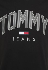 Czarna bawełniana koszulka z wyraźnym białym i szarym napisem "TOMMY" oraz małym symbolem flagi w kolorach czerwonym, białym i niebieskim nad słowem "JEANS."