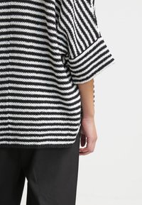 Personne portant un pull rayé noir et blanc à l'horizontale et un pantalon noir, vue de dos, bras relâché le long du corps.