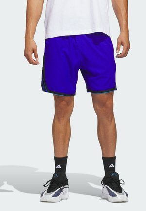 Mężczyzna stoi, ubrany w jasnoniebieskie sportowe szorty, białe buty sportowe z czarnymi i niebieskimi detalami oraz czarne skarpetki Adidas.