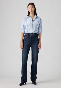 Sieviete stāv tumši zilās augstās džinsās, gaiši zilā svītrainā krekliņā ar uzrullētām piedurknēm un melnās garās kurpēs uz pelēka fona.