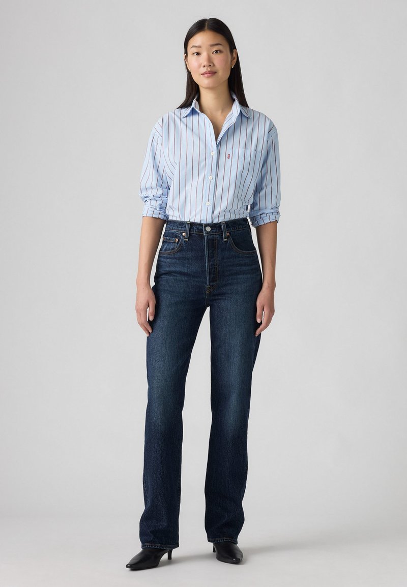 Žena stoji u tamnoplavo visokim trapericama, svijetloplavoj košulji s prugama i zavinutim rukavima, te crnim cipelama s oštrim vrhom na sivoj pozadini.