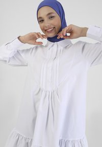 Biała bluzka z długim rękawem, zapinana na guziki, z plisowanym wzorem i marszczonym dołem. Modelka ma na sobie niebieski hijab.