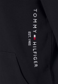 Schwarzer Sweatshirt mit weißem, gesticktem Text "TOMMY HILFIGER EST. 1985" und einem roten und weißen Logo-Akzent. Weicher, atmungsaktiver Stoff.