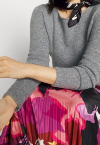 Grijs gebreide trui met lange mouwen gecombineerd met een geplooide, meerkleurige rok met gedurfde roze en rode bloemenpatronen.