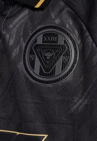 Svart sportströja med texturerad tyg, en cirkulär svart emblem med en triangel och året "XXIII", samt guld accenter längs kragen.