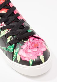 Basket noire avec lacets noirs ornée de grands motifs floraux roses et de feuilles vertes sur l’avant-pied et les côtés, semelle blanche visible.