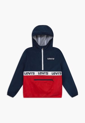 Röd och marinblå vindjacka med halvdragkedja, huva, framficka och vit logotyp i midjan. Tillverkad av lättviktig tyg.