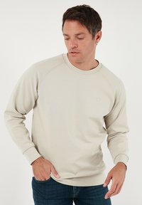 Sudadera beige claro con cuello redondo y mangas raglán. Tela suave, detalle de logo minimalista y puños acanalados. Combinada con vaqueros azules.