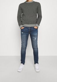 Grauer Strickpullover mit einem helleren grauen Rundhalsausschnitt, kombiniert mit ausgewaschenen blauen Skinny-Jeans und weißen Sportschuhen.