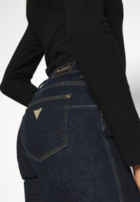 Mörkblå denimjeans med gula sömmar, som har en triangulär logotypapplikation på bakdelen av midjan och två bakfickor. Strukturerad tyg.