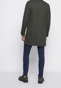 Manteau long vert foncé à coupe droite, tissu texturé, et poignets boutonnés, porté avec un jean slim bleu foncé et des baskets noires.