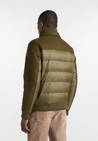 Giacca imbottita di colore verde oliva con una parte superiore simile alla lana e sezioni inferiori lisce e trapuntate. Presenta un colletto alto e polsini a costine.