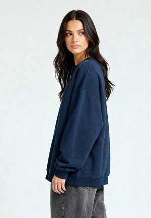 Kvinna med långt mörkt hår som bär en lös navyblå sweatshirt och grå jeans, stående på sidan och tittar åt vänster.