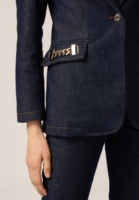 Blazer in denim di un blu navy scuro con un accento in catena dorata sulla tasca laterale. Presenta un design sartoriale, cuciture arancioni a contrasto e una silhouette strutturata.