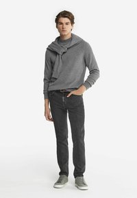 Grau gestrickter Pullover mit einem Schalkragen, kombiniert mit dunkelgrauen Skinny-Jeans und hellgrauen Turnschuhen, vor einem weißen Hintergrund stehend.