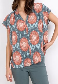 Kortærmet bluse i slub-stof, med en blå baggrund og orange samt lyseblå blomster mønstre og en v-halsudskæring.