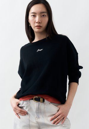 Pitkähiuksinen nainen, jolla on musta neule, jossa valkoista tekstiä, alla punainen paita, vaaleat raidalliset housut ja musta vyö, jossa kullanvärinen solki.