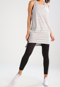 Tunic senza maniche a righe grigie con orlo curvo, indossata su leggings neri con strisce laterali bianche, abbinata a scarpe sportive bianche.
