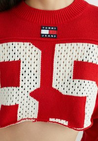 Červený zkrácený pletený svetr s velkým bílým číslem "35" a síťovým vzorem. Etiketa zní "TOMMY JEANS" v modré a bílé barvě.