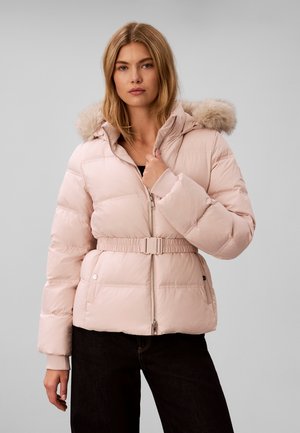 Femme portant une veste matelassée rose clair avec ceinture et capuche bordée de fourrure, debout devant un fond gris uni.