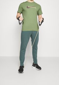 Camiseta de algodón verde con el texto "Entrenamiento", pantalones de chándal verdes y zapatillas negras, sosteniendo bandas de resistencia con mangos negros.