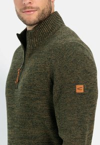 Grüner Strickpullover mit geripptem Kragen und halbem Reißverschluss, ausgestattet mit einem Lederlogo-Patch am Ärmel und einem strukturierten Stoffmuster.
